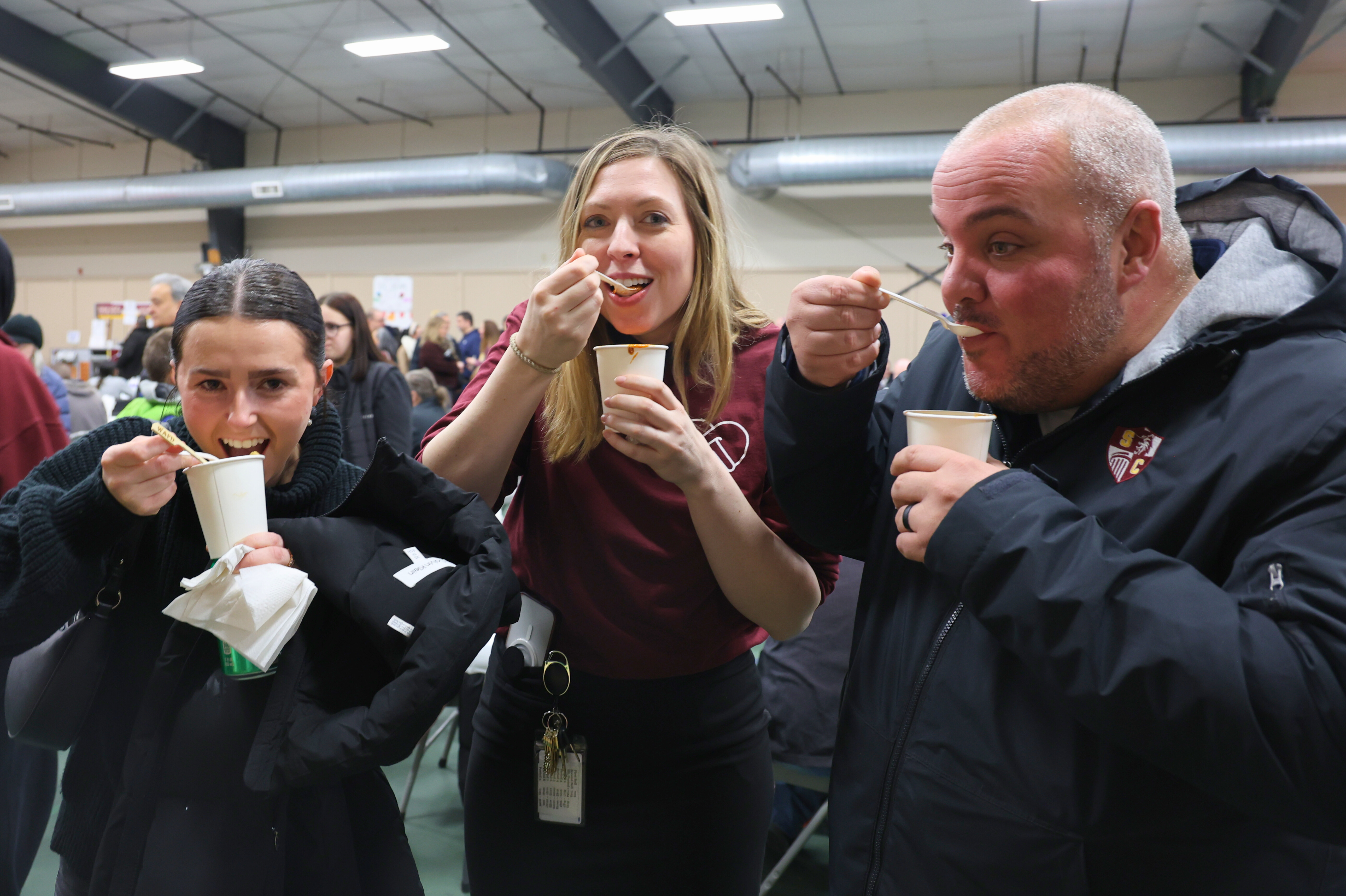 three adults hold white cups up while they take a bite of chili in a crowded room